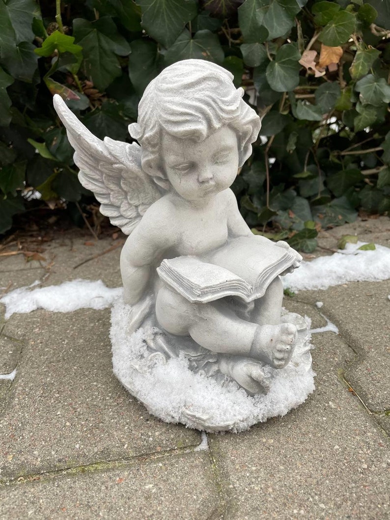 May include: A gray stone cherub angel statue with wings, sitting on a snowy base, holding an open book. The angel has a sweet expression on its face.
