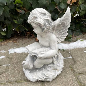 May include: A gray stone sculpture of an angel with wings, sitting on a rock and reading a book.