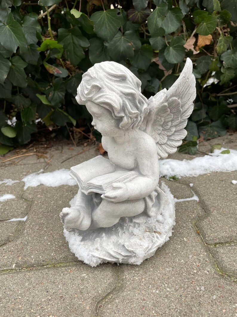May include: A gray stone sculpture of an angel with wings, sitting on a rock and reading a book.