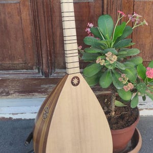Puede incluir: Un instrumento musical de cuerda conocido como saz o baglama, con un cuerpo de madera clara en forma de pera, un m&aacute;stil largo con trastes finos y una incrustaci&oacute;n decorativa en forma de estrella en la tapa, sobre un soporte de metal.