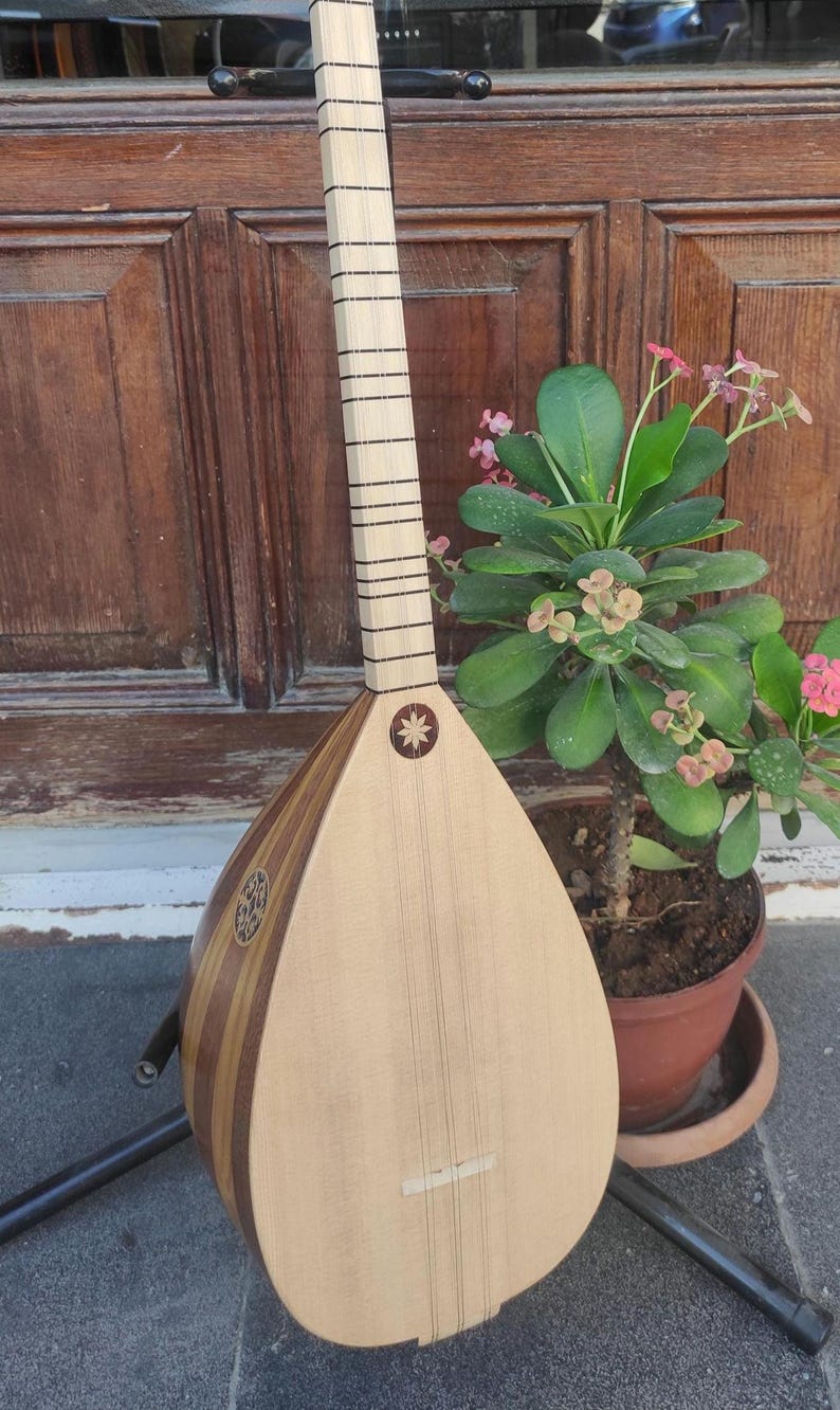 Puede incluir: Un instrumento musical de cuerda conocido como saz o baglama, con un cuerpo de madera clara en forma de pera, un m&aacute;stil largo con trastes finos y una incrustaci&oacute;n decorativa en forma de estrella en la tapa, sobre un soporte de metal.