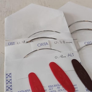 Puede incluir: Sobres de papel blanco que contienen agujas met&aacute;licas curvas para instrumentos musicales. Los sobres tienen etiquetas con texto que incluye 0,18 mm y 0,28 mm, y cuentan con leng&uuml;etas ovaladas de pl&aacute;stico rojo y marr&oacute;n oscuro. Texto: BAĞLAMA, ALT, ORTA, &Uuml;ST.