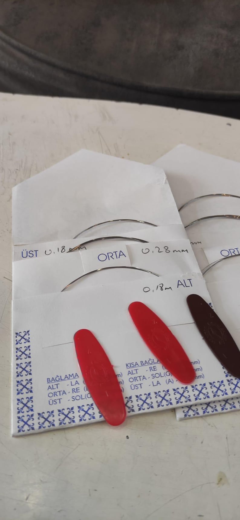 Puede incluir: Sobres de papel blanco que contienen agujas met&aacute;licas curvas para instrumentos musicales. Los sobres tienen etiquetas con texto que incluye 0,18 mm y 0,28 mm, y cuentan con leng&uuml;etas ovaladas de pl&aacute;stico rojo y marr&oacute;n oscuro. Texto: BAĞLAMA, ALT, ORTA, &Uuml;ST.