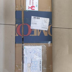 Puede incluir: Una caja de env&iacute;o rectangular y alta de cart&oacute;n envuelta en cinta adhesiva transparente. La caja presenta una banda horizontal azul con el texto OMEK, una etiqueta blanca de fr&aacute;gil con texto en rojo, una etiqueta de env&iacute;o y una funda pl&aacute;stica.