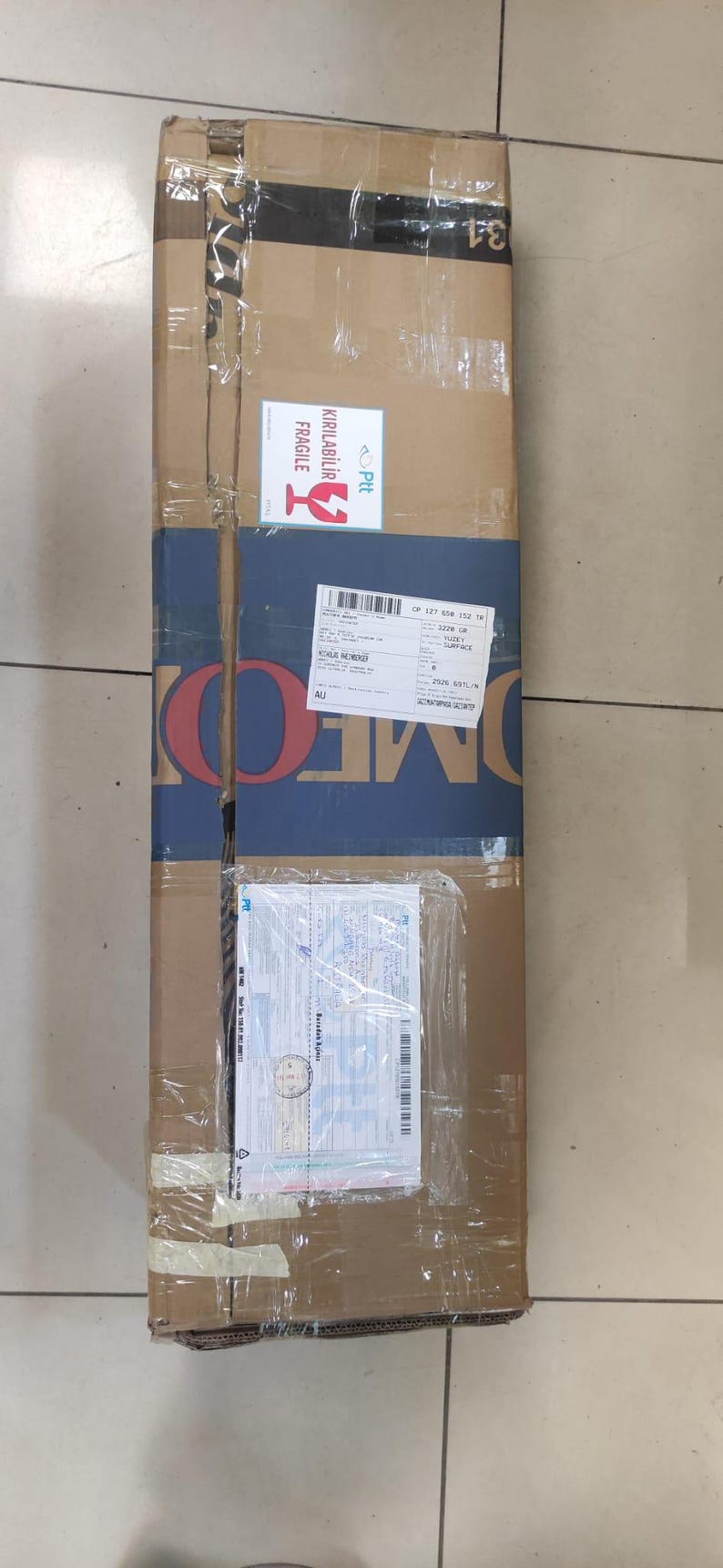 Puede incluir: Una caja de env&iacute;o rectangular y alta de cart&oacute;n envuelta en cinta adhesiva transparente. La caja presenta una banda horizontal azul con el texto OMEK, una etiqueta blanca de fr&aacute;gil con texto en rojo, una etiqueta de env&iacute;o y una funda pl&aacute;stica.