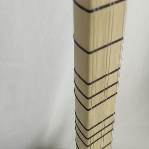 Puede incluir: Un m&aacute;stil de madera de color claro de un instrumento musical de cuerda, con m&uacute;ltiples trastes finos y oscuros en forma de V y varias cuerdas verticales tensas. El m&aacute;stil se conecta a un cuerpo de madera redondeado y de tonos variados con una boca.
