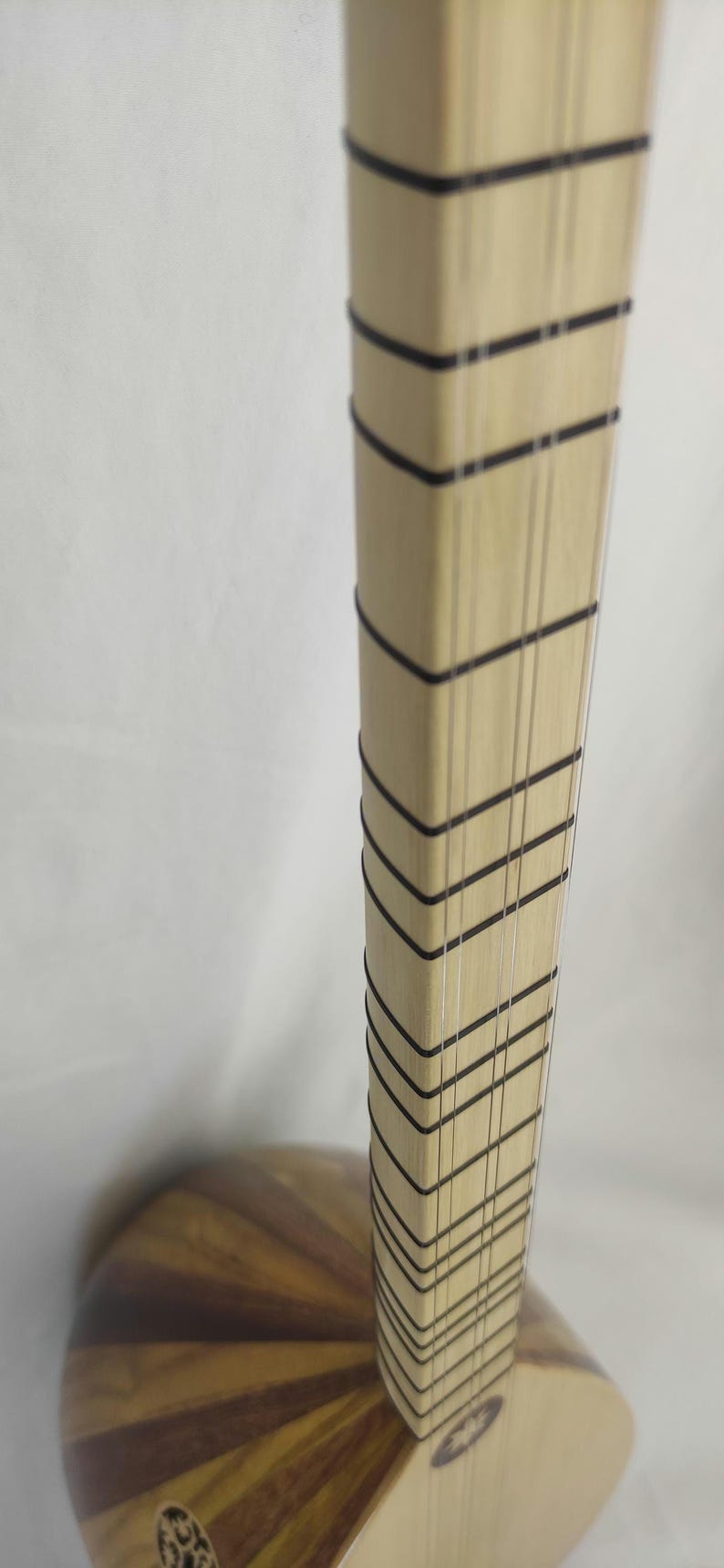 Puede incluir: Un m&aacute;stil de madera de color claro de un instrumento musical de cuerda, con m&uacute;ltiples trastes finos y oscuros en forma de V y varias cuerdas verticales tensas. El m&aacute;stil se conecta a un cuerpo de madera redondeado y de tonos variados con una boca.