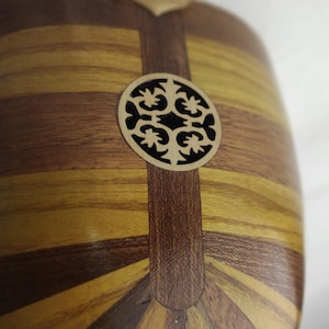 Puede incluir: Un cuerpo de instrumento musical de madera redondeado con paneles de vetas de madera alternas en marr&oacute;n claro y oscuro. Una incrustaci&oacute;n circular de madera clara con un patr&oacute;n de corte sim&eacute;trico e intrincado est&aacute; centrada.