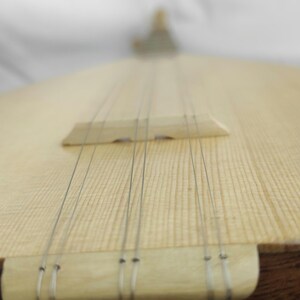 Puede incluir: Un instrumento musical de cuerda con una caja de resonancia de madera clara y veta visible. Seis cuerdas met&aacute;licas finas recorren verticalmente el cuerpo, pasando sobre un puente de madera y ancladas en un cordal de madera clara.
