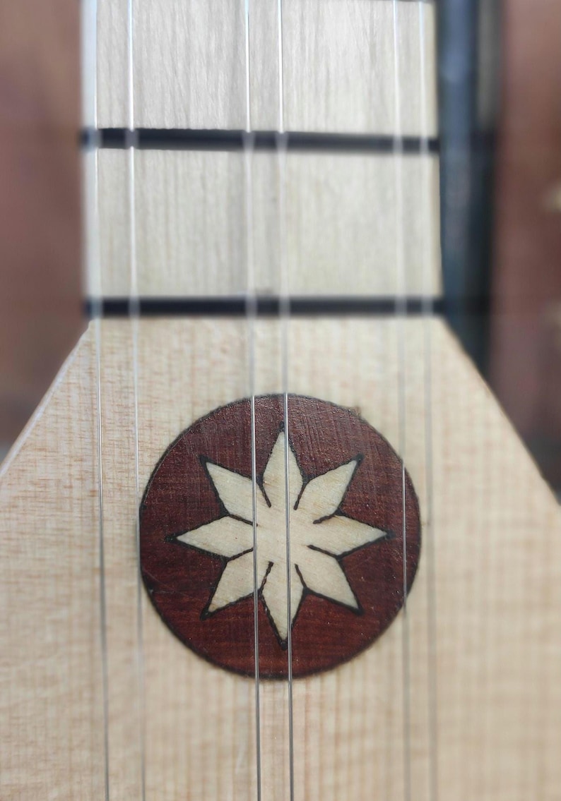 May include: A light-colored wooden stringed instrument neck featuring a circular inlay with a dark brown background and a light-colored eight-pointed star design. Four thin metal strings run vertically across the fretboard and the inlay.