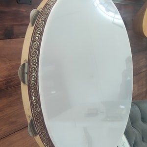 May include: A white tambourine with a gold and brown decorative border. The tambourine has a wooden frame and metal jingles.