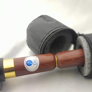 Puede incluir: Una herramienta de masaje de madera con un extremo de goma negra y detalles dorados, parcialmente encerrada en un estuche negro con cremallera. La herramienta tiene un mango de madera marrón con una banda dorada y una pegatina azul y blanca que dice "Do".