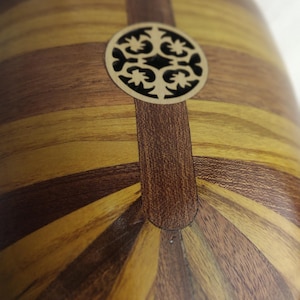 Puede incluir: Un cuerpo de instrumento musical de madera con una parte posterior redondeada en forma de cuenco, construida con duelas de madera alternas en marr&oacute;n oscuro y bronceado claro. Un roset&oacute;n circular de madera cortado con l&aacute;ser y dise&ntilde;o floral.