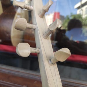 Puede incluir: Un clavijero de instrumento musical de madera clara con seis clavijas de afinaci&oacute;n dispuestas en dos filas. Las clavijas son de madera p&aacute;lida y lisa, y est&aacute;n conectadas a finas cuerdas met&aacute;licas que recorren el m&aacute;stil del instrumento.