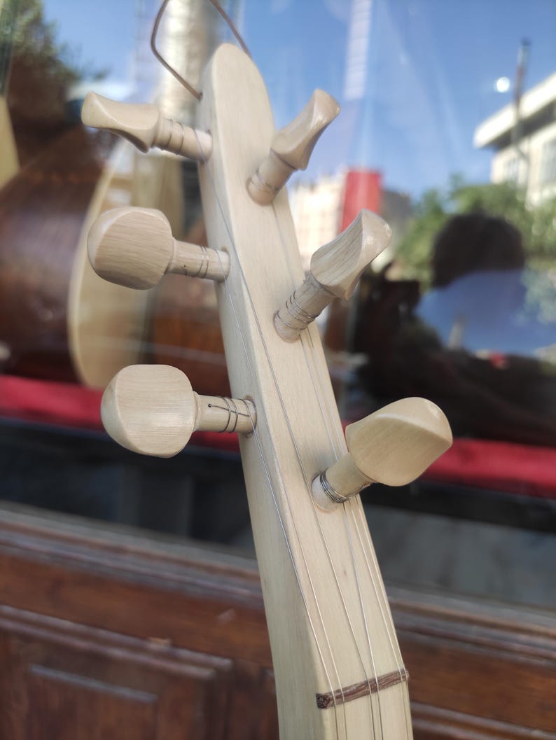 Puede incluir: Un clavijero de instrumento musical de madera clara con seis clavijas de afinaci&oacute;n dispuestas en dos filas. Las clavijas son de madera p&aacute;lida y lisa, y est&aacute;n conectadas a finas cuerdas met&aacute;licas que recorren el m&aacute;stil del instrumento.