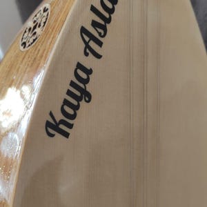 Puede incluir: Un instrumento de cuerda saz de madera clara con cuerpo en forma de pera, que presenta el nombre Kaya Aslan impreso en letras negras sobre la tapa arm&oacute;nica, cuerdas met&aacute;licas finas, un puente blanco y una incrustaci&oacute;n decorativa.
