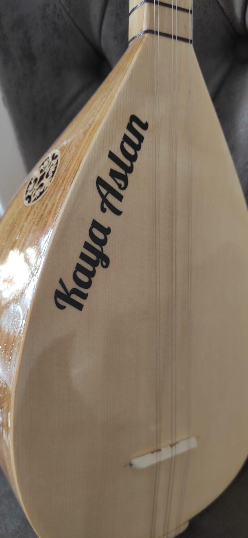 Puede incluir: Un instrumento de cuerda saz de madera clara con cuerpo en forma de pera, que presenta el nombre Kaya Aslan impreso en letras negras sobre la tapa arm&oacute;nica, cuerdas met&aacute;licas finas, un puente blanco y una incrustaci&oacute;n decorativa.