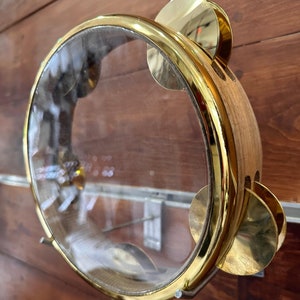 Op de afbeelding: Een gouden tamboerijn met een doorzichtig plastic vel en messing schellen. De tamboerijn is gemaakt van hout en heeft een ronde vorm.