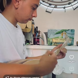 Puede incluir: Una persona tocando un instrumento de cuerda de madera conocido como cura, con un cuerpo redondeado y un m&aacute;stil largo con trastes. El instrumento se sostiene horizontalmente mientras la persona pulsa las cuerdas y toca el m&aacute;stil.