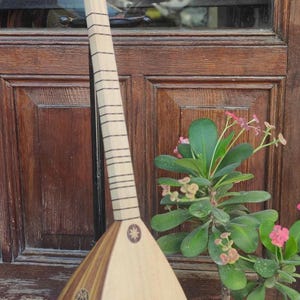 Puede incluir: Un instrumento de cuerda de m&aacute;stil largo con un cuerpo de madera en forma de pera que presenta paneles de vetas de madera clara y oscura, un m&aacute;stil delgado con m&uacute;ltiples trastes y clavijas de afinaci&oacute;n claras, sobre un soporte de metal negro.