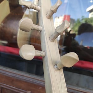Puede incluir: Un clavijero de madera de color claro de un instrumento musical de cuerda con seis clavijas de afinaci&oacute;n dispuestas en tres pares a los lados. Se aprecia la veta de la madera lisa y p&aacute;lida, con cuerdas finas sujetas a las clavijas.