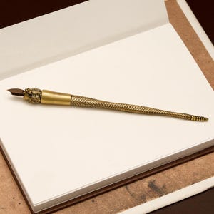 May include: A brass dip pen with a snake-scale design and a serpent head detail. The pen is displayed on a white notepad, highlighting its intricate design and vintage aesthetic. The nib is dark brown.