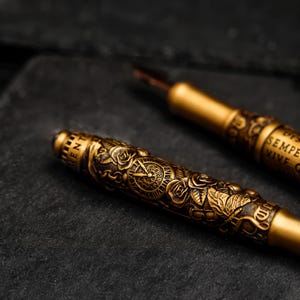 May include: Two gold-toned fountain pens with intricate designs. One pen features a clock face and roses, while the other has the words "SEMPER VIVE" engraved on it.