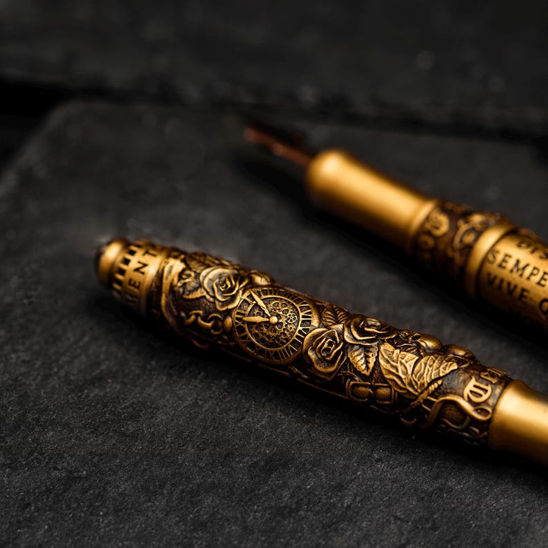 May include: Two gold-toned fountain pens with intricate designs. One pen features a clock face and roses, while the other has the words "SEMPER VIVE" engraved on it.