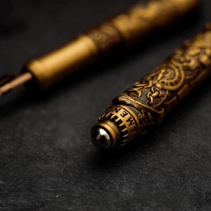 May include: Close-up of a gold-toned pen with intricate engraved details. The pen has a round, silver-toned tip and the word "MEN" is visible on the side.