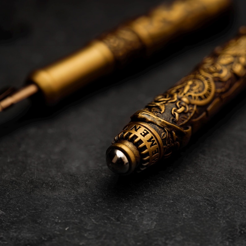 May include: Close-up of a gold-toned pen with intricate engraved details. The pen has a round, silver-toned tip and the word "MEN" is visible on the side.