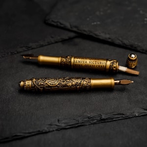 May include: Two ornate gold-toned fountain pens with intricate designs and inscriptions. The inscription on one pen reads "Disce Quasi Semper Victorus Vive Quasi Cras".
