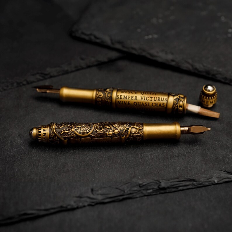 May include: Two ornate gold-toned fountain pens with intricate designs and inscriptions. The inscription on one pen reads "Disce Quasi Semper Victorus Vive Quasi Cras".
