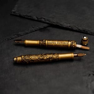 May include: Two gold-toned fountain pens with intricate designs and a Latin inscription: "Disce Quasi Semper Victorus Vive Quasi Cras". One pen is open, revealing a nib and a small, round, silver-colored cap.