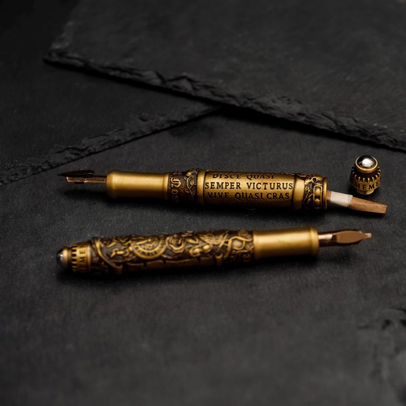May include: Two gold-toned fountain pens with intricate designs and a Latin inscription: "Disce Quasi Semper Victorus Vive Quasi Cras". One pen is open, revealing a nib and a small, round, silver-colored cap.