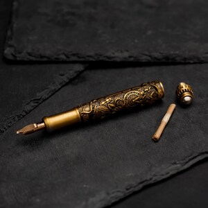 May include: A gold-toned fountain pen with an intricate floral design and a separate cap. The pen is lying on a black surface.