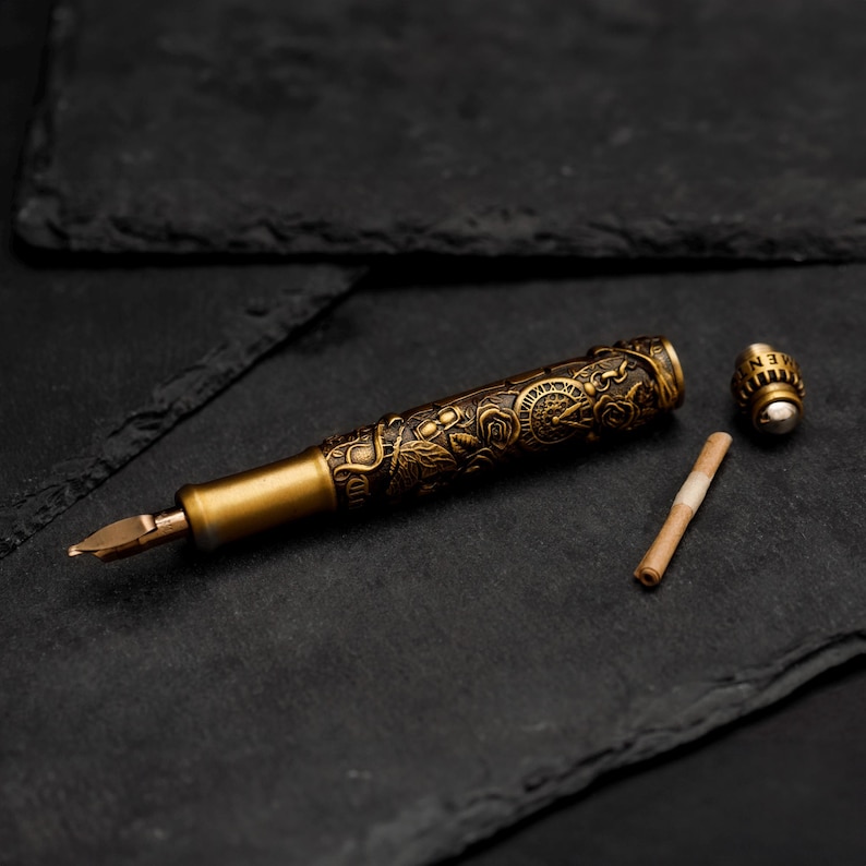 May include: A gold-toned fountain pen with an intricate floral design and a separate cap. The pen is lying on a black surface.