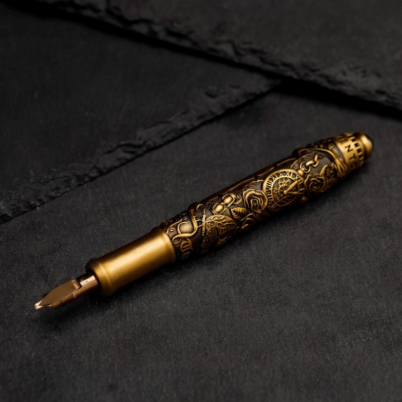 May include: A gold-toned fountain pen with an intricate design featuring a clock, flowers, and vines. The pen has a fine nib and a cap with the word "INTENT" engraved on it.