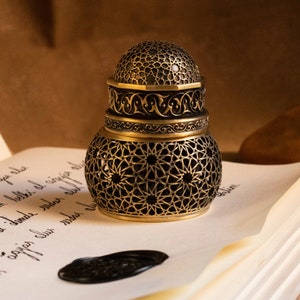 Peut inclure: Un encrier en laiton antique avec des motifs géométriques et floraux découpés. L'encrier est posé sur une feuille de papier avec une écriture manuscrite et un sceau de cire noir. Le couvercle de l'encrier est en forme de dôme.