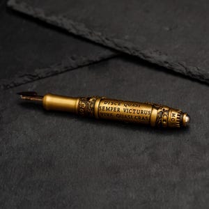 May include: A gold-toned pen with an ornate design and a nib at one end. The pen has an inscription that reads "DISCE QUASI SEMPER VICTURUS VIVE QUASI CRAS".