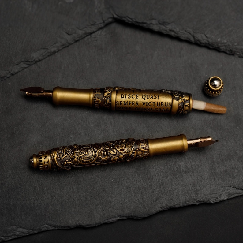 May include: Two gold-toned fountain pens with intricate designs and inscriptions. The top pen has the inscription "DISCE QUASI SEMPER VICTURUS".