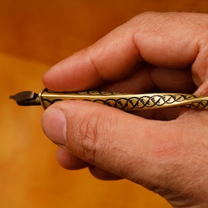 Peut inclure: Un stylo de calligraphie en métal doré doté d'un corps torsadé en spirale avec un motif gravé de couleur foncée complexe le long de sa longueur et une plume en métal pointue à l'extrémité pour écrire ou dessiner.