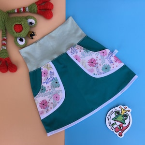 May include: A teal skirt with white trim and floral fabric pockets. The skirt has a light green waistband.