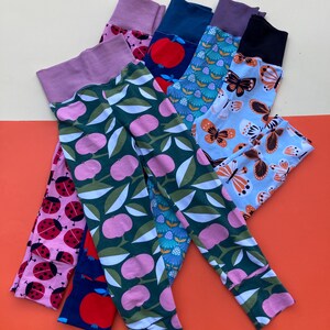 Peut inclure: Cinq paires de leggings colorés avec différents motifs empilés. Les motifs comprennent des coccinelles, des pommes, des fleurs et des papillons. Les leggings sont faits d'un tissu doux et extensible.