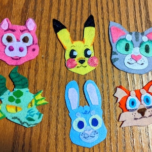 May include: A collection of six colorful felt animal face ornaments. The ornaments include a pink pig, a yellow Pikachu, a gray cat, a green dragon, a blue bunny, and an orange fox. Each ornament is hand-stitched with detailed features.