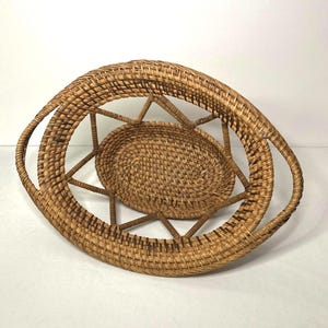 May include: Woven rattan basket with a unique oval shape and integrated handles. The basket features a central circular base and a starburst design connecting the base to the oval rim. The natural brown color of the rattan gives it a rustic aesthetic.