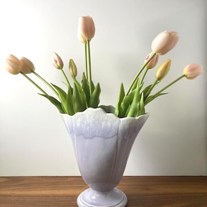 May include: A light purple vase with a scalloped rim holds several pink tulips with green stems and leaves. The vase has a pedestal base and is set on a wooden surface. The tulips are in various stages of bloom.