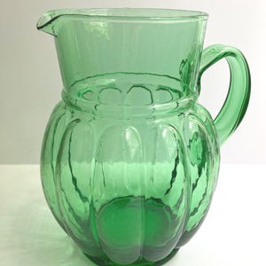 May include: A translucent emerald green glass pitcher featuring a rounded, ribbed body with vertical indentations, a wide circular rim with a pouring spout, and a curved handle attached to the side of the vessel for easy gripping and pouring.
