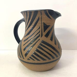 May include: A ceramic pitcher with a black handle and spout. The pitcher has a tan base color with a black geometric pattern. The design features angled lines and triangles. The pitcher is a decorative item.