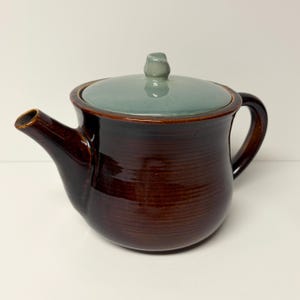 May include: A ceramic teapot with a glossy dark brown body featuring horizontal ribbed textures, a curved handle, and a short spout. The teapot includes a removable lid with a smooth, light sage green glaze and a small central knob handle.