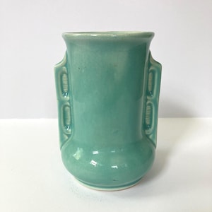 May include: A teal-colored ceramic vase with a glossy finish. The vase has a rounded base, a cylindrical body, and a flared rim. Two decorative, rectangular handles are on either side of the vase. The vase is a solid color with no other patterns or designs.